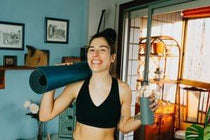 Smiling woman in black sports bra carrying a blue yoga mat and water bottle indoors.