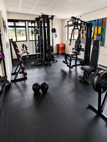 A spacious home gym interior with black rubber flooring, featuring multiple pieces of fitness equipment including a cable machine, weight bench, dumbbell rack, and medicine balls.